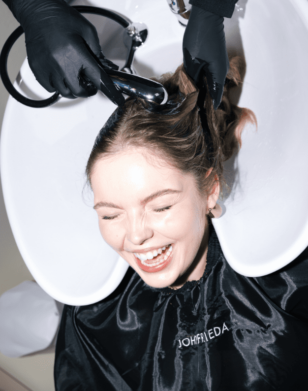 Smiling woman enjoying a salon hair wash while seated at a shampoo bowl, with a stylist using a handheld showerhead to rinse her hair, wearing a black cape.