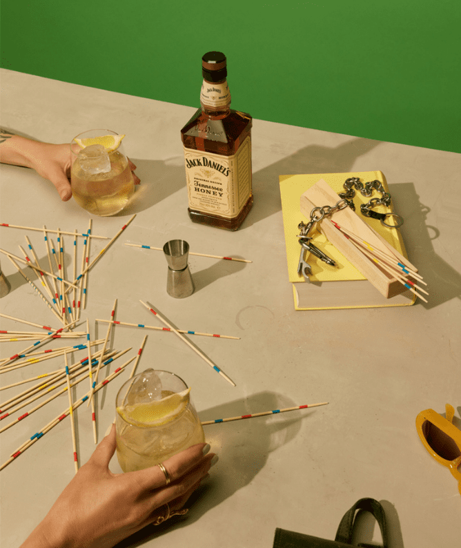 2 A top-down view of a tabletop scene featuring a hand holding a cocktail with ice and a lemon slice, a bottle of Jack Daniel’s Tennessee Honey, and various game pieces including colorful toothpicks scattered across a light-colored surface. Nearby, a measuring jigger, a yellow notebook, and a set of keys are also visible, creating a vibrant and engaging atmosphere for a social gathering.