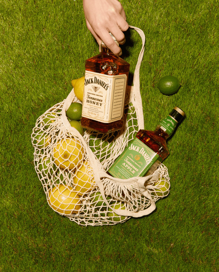 3 Bottles of Jack Daniel's Tennessee Honey and Apple whiskey partially nestled in a net bag filled with fresh lemons and limes, set against a vibrant green grass backdrop.