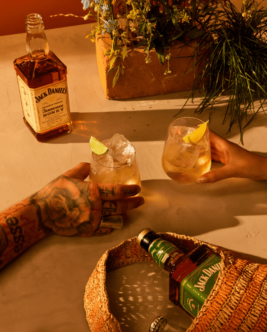 4 Two glasses of mixed drinks with lime and lemon garnishes are being held, accompanied by a bottle of Jack Daniel's Tennessee Honey and another bottle in the foreground, set against a warm, inviting background with greenery.