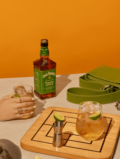 5 A bottle of Jack Daniel's Tennessee Apple whiskey is placed on a table alongside two cocktail glasses filled with ice and garnished with lime. A green handbag and a jigger sit next to the drinks against a vibrant orange background.