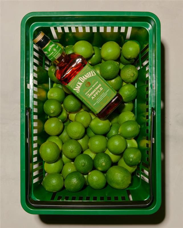 JD 1 Basket filled with fresh green limes and a bottle of Jack Daniel's Tennessee Apple whiskey, showcasing a vibrant combination ideal for cocktails and summer drinks.