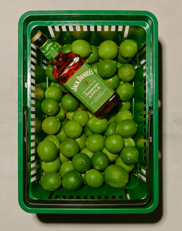 Basket filled with vibrant green limes and a bottle of Jack Daniel's Tennessee Apple whiskey, showcasing a fresh and fruity beverage theme.