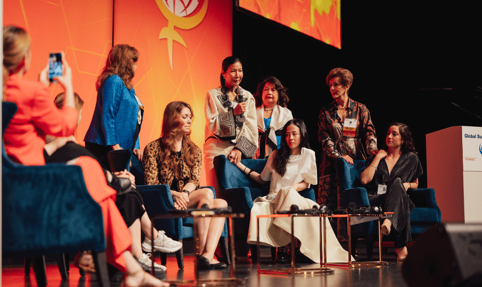 news_GSOW A diverse group of women speakers engaging in discussion on stage at the Global Summit, showcasing themes of empowerment and collaboration, with vibrant backdrop featuring the women's symbol.