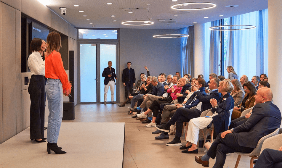 news_luxurybusinessday Two women are presenting on stage during a corporate event, engaging with an audience seated in a modern meeting space. The audience, comprised of diverse professionals, shows interest and interaction while the presenters deliver their talk. The room features contemporary design elements like circular lighting and large windows.