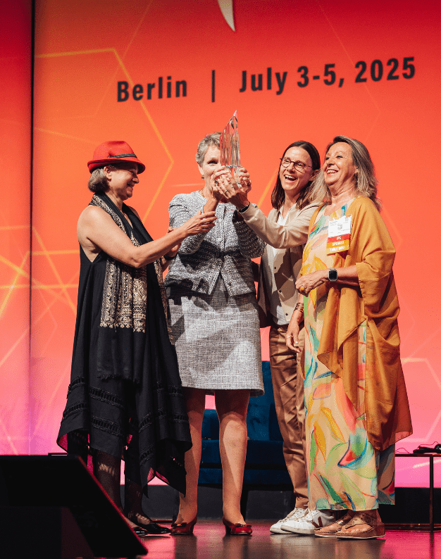 Four women celebrating on stage at a conference in Berlin, July 3-5, 2025, as one of them holds an award. They are dressed in diverse outfits, showcasing joy and achievement against a vibrant background.