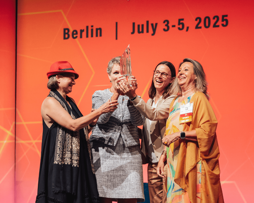 2 Four women celebrate on stage in Berlin, July 2025, as they jointly hold an award, showcasing a moment of achievement and camaraderie during the event. The background features vibrant colors and event details.