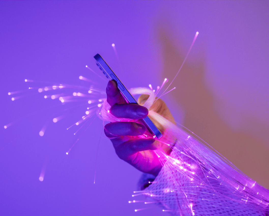 A hand holding a smartphone is surrounded by glowing fiber optic strands, illuminated in a purple hue, symbolizing technology and connectivity.