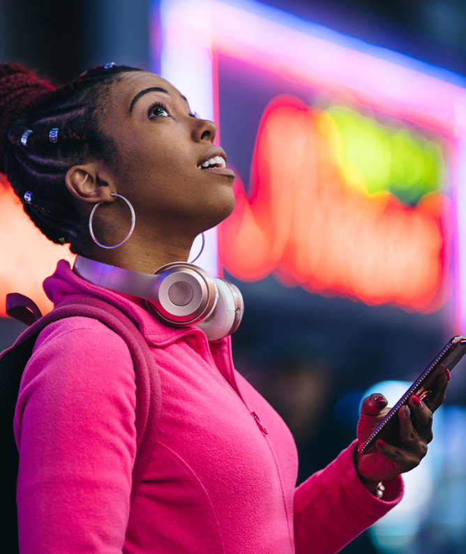 Young woman in a pink fleece jacket wearing headphones, looking up with a smartphone in her hand against a colorful neon backdrop.