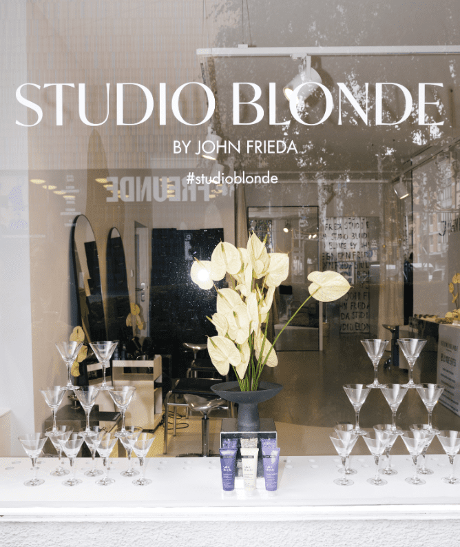 3 Window display of Studio Blonde by John Frieda featuring elegant floral arrangements, hair products, and a stylish interior. Showcases a modern salon atmosphere with glassware and salon chairs visible through the glass.