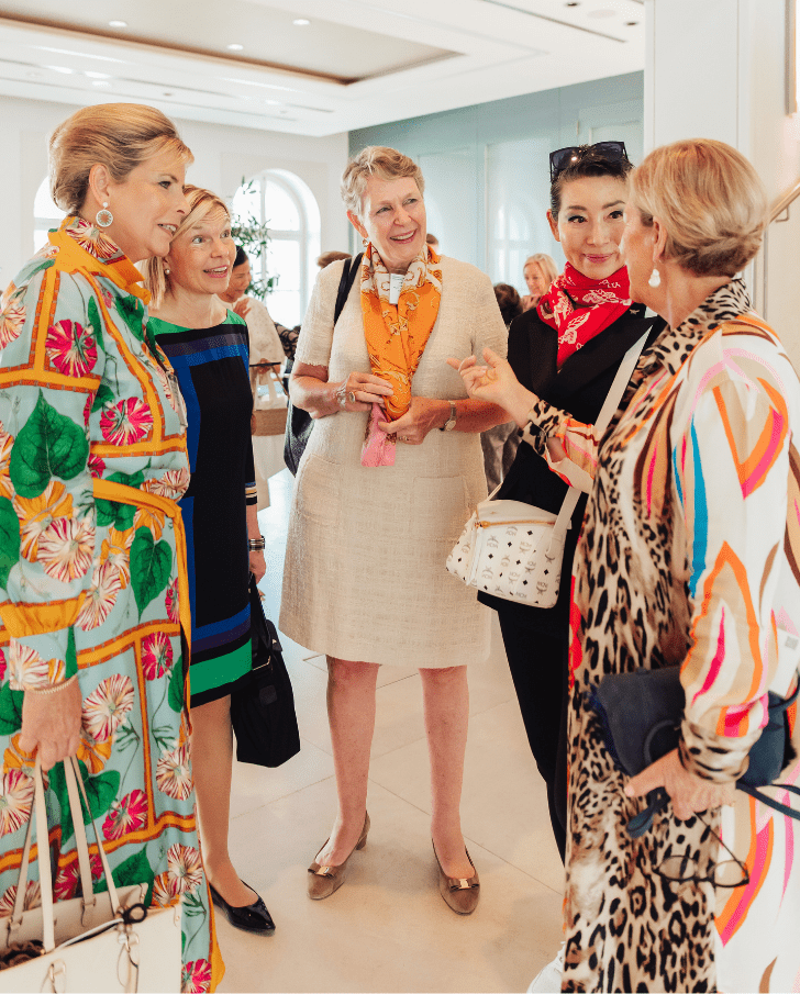 4 A group of five stylish women engaged in conversation at a social event, showcasing vibrant fashion choices and accessories, set in a bright and elegant interior.