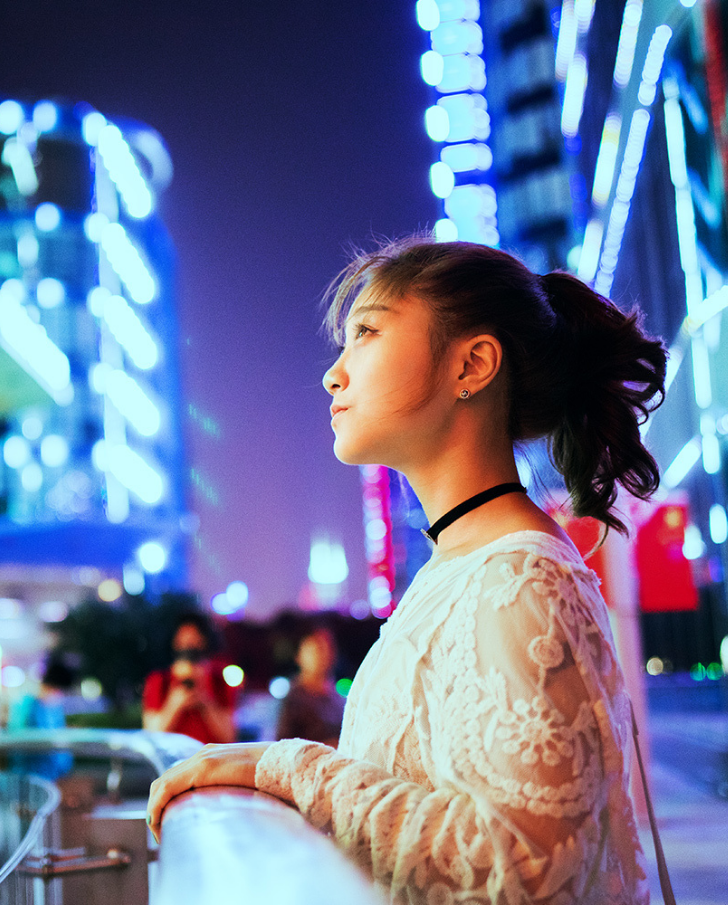 Young woman in a stylish lace top gazing thoughtfully at a vibrant cityscape illuminated by colorful lights at night. The background features modern architecture and urban scenery, highlighting the dynamic atmosphere of the city.