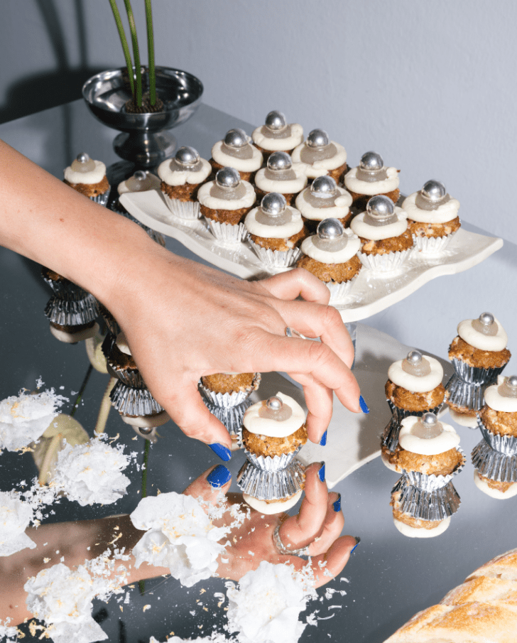 4 A hand reaching for decorative cupcakes with silver toppings, arranged on a reflective surface, surrounded by scattered flower petals and a minimalistic plant in a pot.