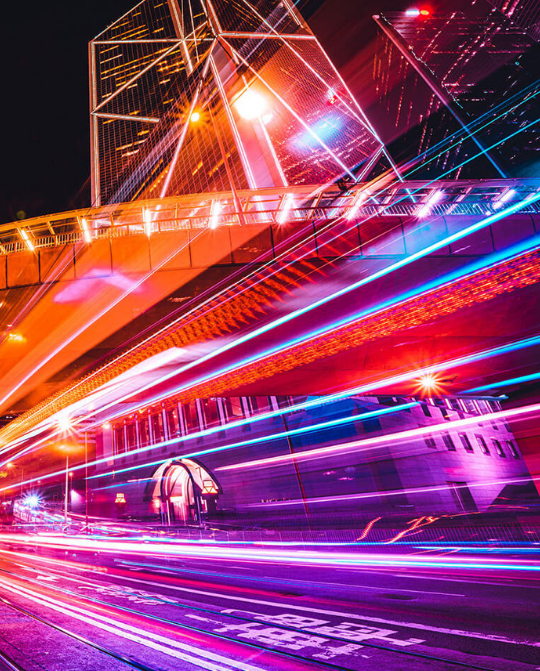 Vibrant night view of a futuristic cityscape featuring illuminated skyscrapers and dynamic light trails from passing vehicles on a busy street. The composition highlights the architectural design and urban energy, creating a stunning visual effect.