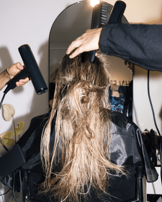 5 A hairstylist using a hairdryer and a straightener on long, unstyled hair in a salon setting, with a mirror reflecting the process and tools.