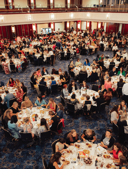 6 Aerial view of a large banquet hall filled with elegantly dressed guests seated at round tables, enjoying a meal during a formal event. The room features stylish decor, and attendees are engaged in conversation while enjoying fine dining.