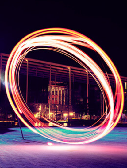 Long exposure photograph of a vibrant, swirling light trail creating a circular pattern at night, with modern architecture and softly illuminated buildings in the background.