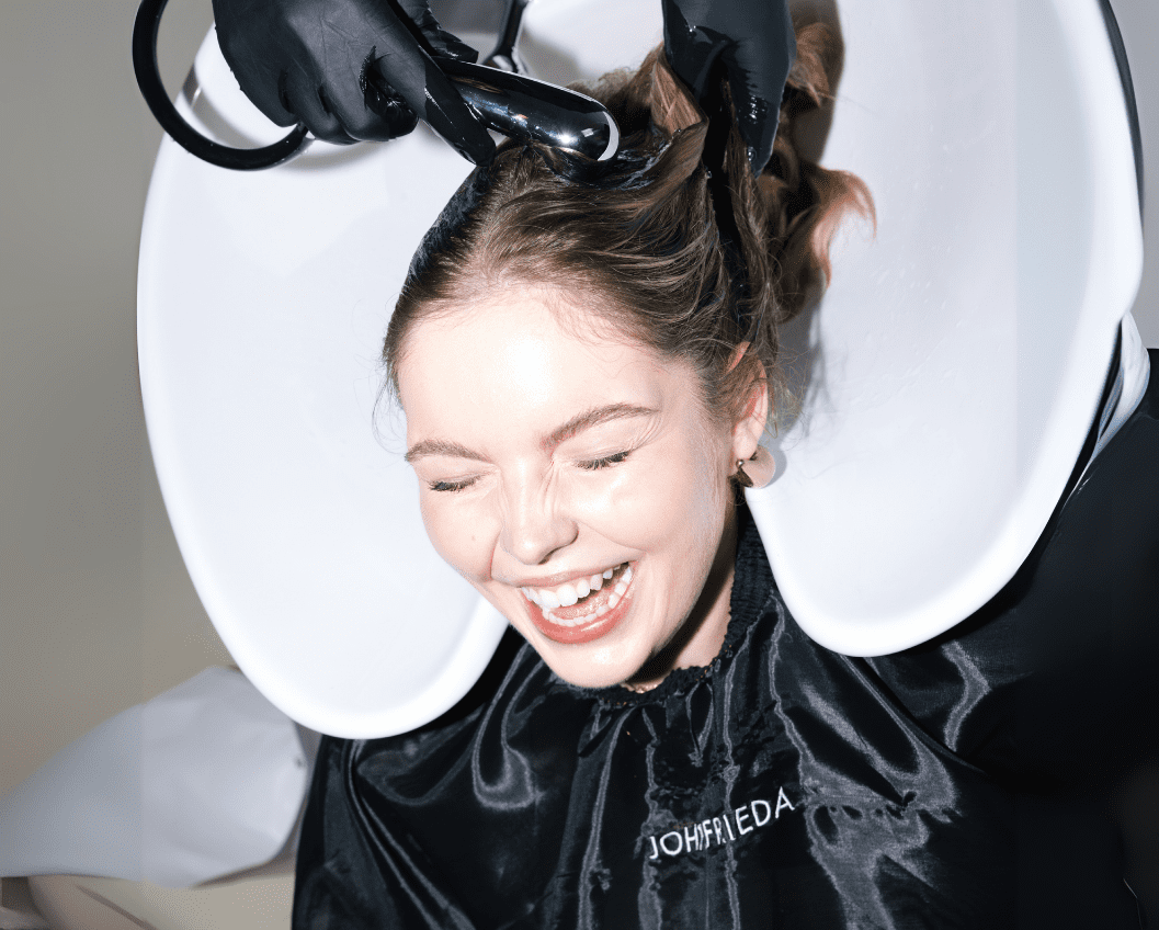 Kopie von VORLAGE Website Cases - Bildgrößen (1) Young woman enjoying a salon shampoo treatment while smiling, seated at a hair washing station with a stylist applying water to her hair, wearing a protective cape.