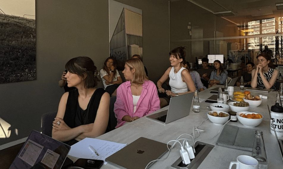12 A diverse group of professionals engaged in a meeting around a conference table, featuring laptops, notepads, and bowls of fruit. The setting showcases a modern office environment with large windows and contemporary decor.