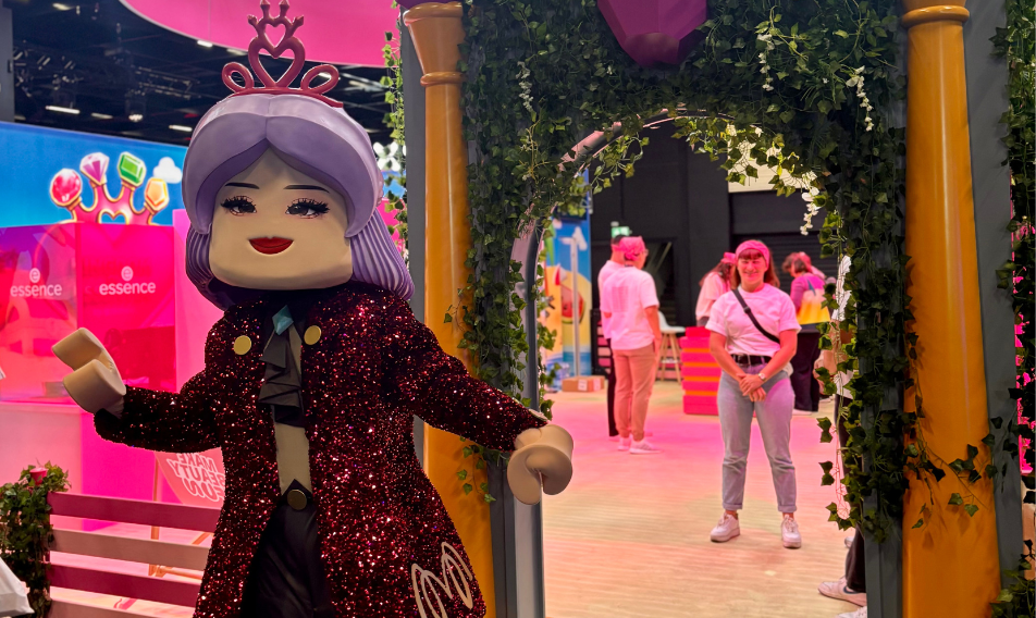 13 Colorful event scene featuring a large, sparkly character figure with purple hair and a crown, posing in front of a decorated archway. In the background, attendees in casual clothing are engaged in activities within a vibrant, playful setting.