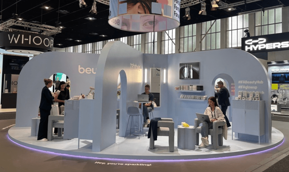 Modern exhibition booth showcasing beauty products, featuring a sleek blue design with interactive displays. Attendees engage with staff, while others work on laptops. The booth emphasizes innovation in the beauty industry, set within a bustling trade show environment.