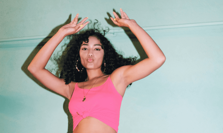 VORLAGE haebmau Website News (4) Young woman with curly hair wearing a pink tank top, dancing against a light green background, showcasing a joyful and energetic expression.