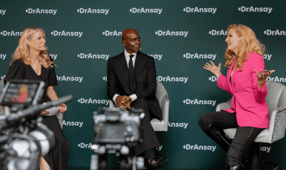Three speakers engage in a discussion during a panel event, with a backdrop featuring the Dr. Ansay logo. The setting includes cameras capturing the conversation, highlighting the dynamic interaction among the presenters.