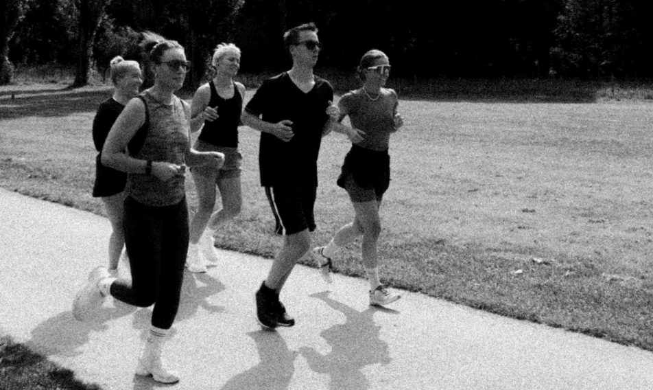 Group of five young adults jogging together on a park path, promoting fitness and outdoor activity. The scene captures a blend of athletic wear and casual styles in a vibrant green landscape.