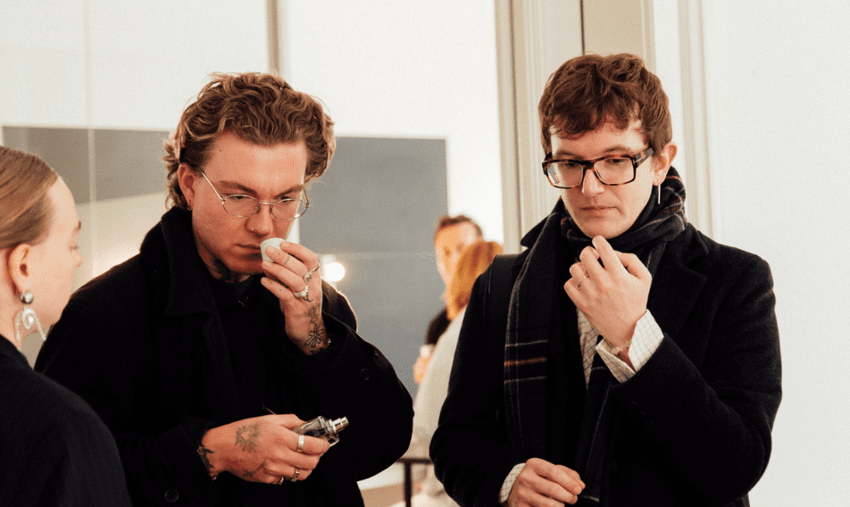Two stylish young men in a modern setting are engaged in a discussion while testing fragrances. One man, wearing glasses and a black overcoat, holds a perfume bottle and is smelling a tester, while the other man, dressed in a scarf and a blazer, observes closely. A woman is partially visible on the left, adding to the social atmosphere of the scene.