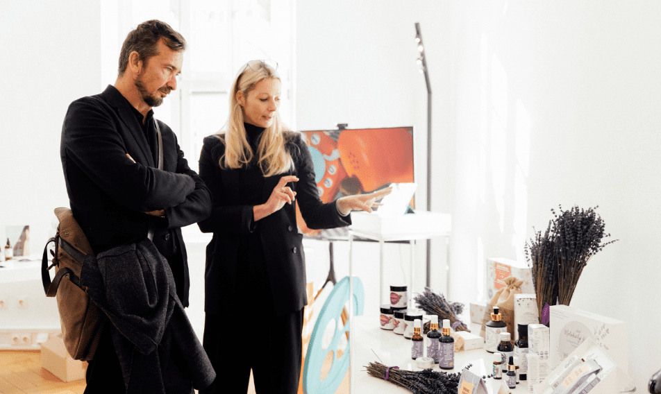 A man and a woman engage in conversation while examining a display of beauty products and lavender in a bright, modern space. The woman gestures towards a tablet, indicating a discussion about the items showcased on the table.