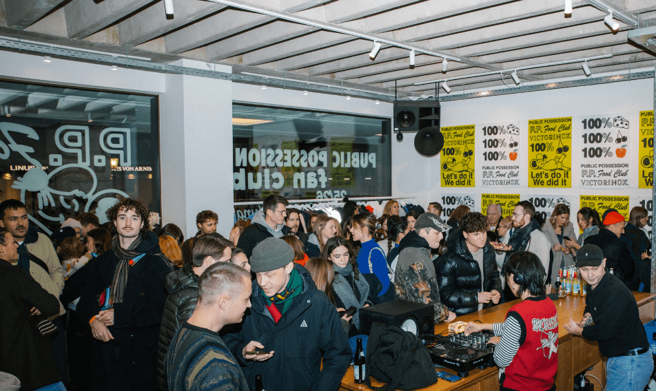 Crowd of people socializing at a vibrant indoor event, featuring a DJ setup and colorful promotional posters on the walls. The atmosphere is lively, with attendees engaging in conversation and enjoying refreshments.