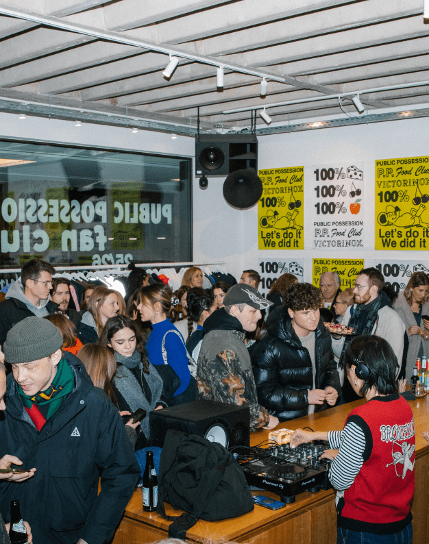 Crowded indoor event at Public Possession featuring a diverse group of attendees socializing, enjoying music from a DJ, and surrounded by vibrant promotional posters.