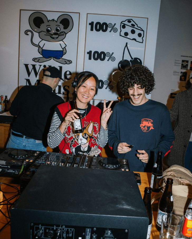 4 Two DJs at a lively event, one woman smiling and holding a drink, and a man with curly hair beside a DJ mixer. Behind them, playful mouse-themed artwork and a display of beverages.
