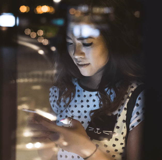A young woman with long hair, wearing a polka dot shirt with a cat design, is focused on her smartphone while illuminated by city lights at night.