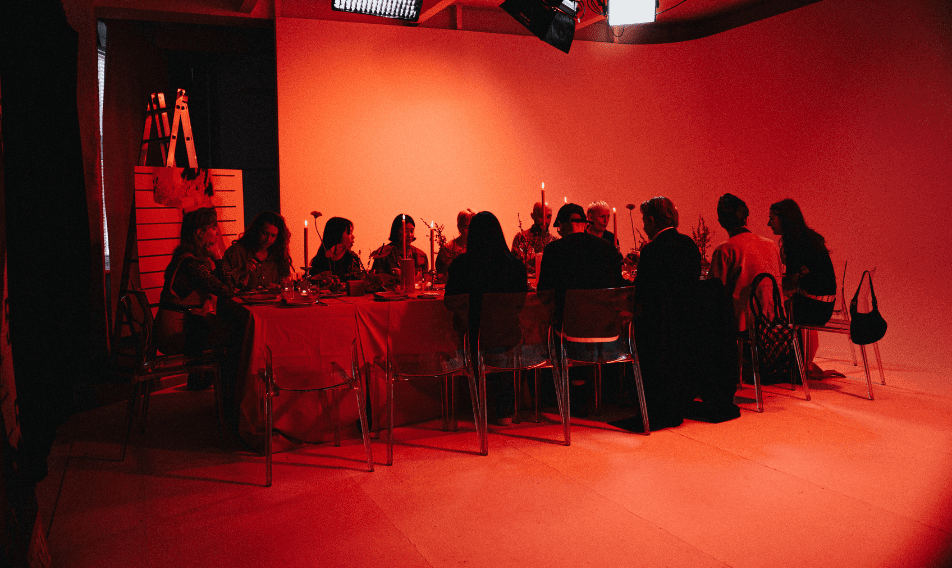 Group of people dining at a long table in a creatively lit studio, featuring red lighting and decorative elements. The scene captures a festive atmosphere with attendees engaged in conversation, set against a minimalist backdrop.