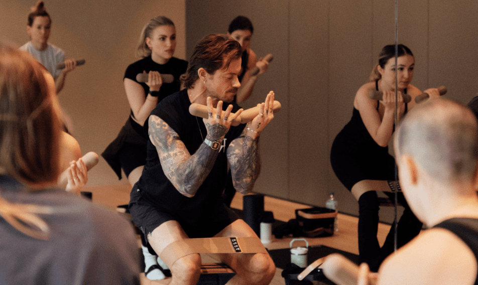 Group fitness class in a studio setting, featuring participants engaged in a workout using small weights and resistance bands. A male instructor demonstrates proper technique while surrounded by focused attendees, showcasing a modern approach to strength training and wellness.